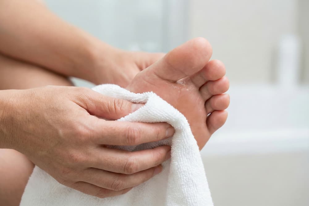 Et nærbillede af hænder, der tørrer fodsålen grundigt med et hvidt håndklæde. Billedet illustrerer vigtigheden af at fjerne fugt fra hele foden efter bad for at forebygge hudlidelser som fodsvamp og bakterievækst.