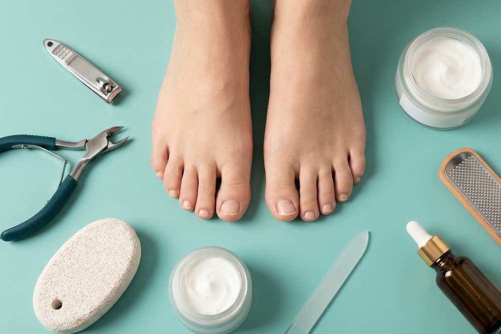 Et organiseret 'flat lay' billede på en turkis baggrund, der viser to bare fødder omgivet af essentielt udstyr til fodpleje. Opstillingen inkluderer negleklippere, en pimpsten til hård hud, fodfile samt krukker med fugtighedscreme og negleolie, hvilket illustrerer de nødvendige redskaber til at opretholde sunde fødder.