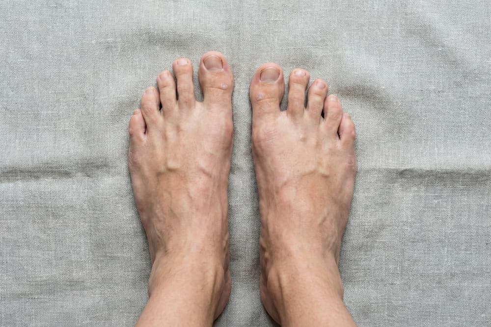 Et billede set oppefra af to bare fødder placeret side om side på en grå stofbaggrund. Billedet illustrerer, at fødder ofte er asymmetriske i størrelse eller form, hvilket understreger vigtigheden af altid at måle og prøve sko på begge fødder for at sikre korrekt pasform.