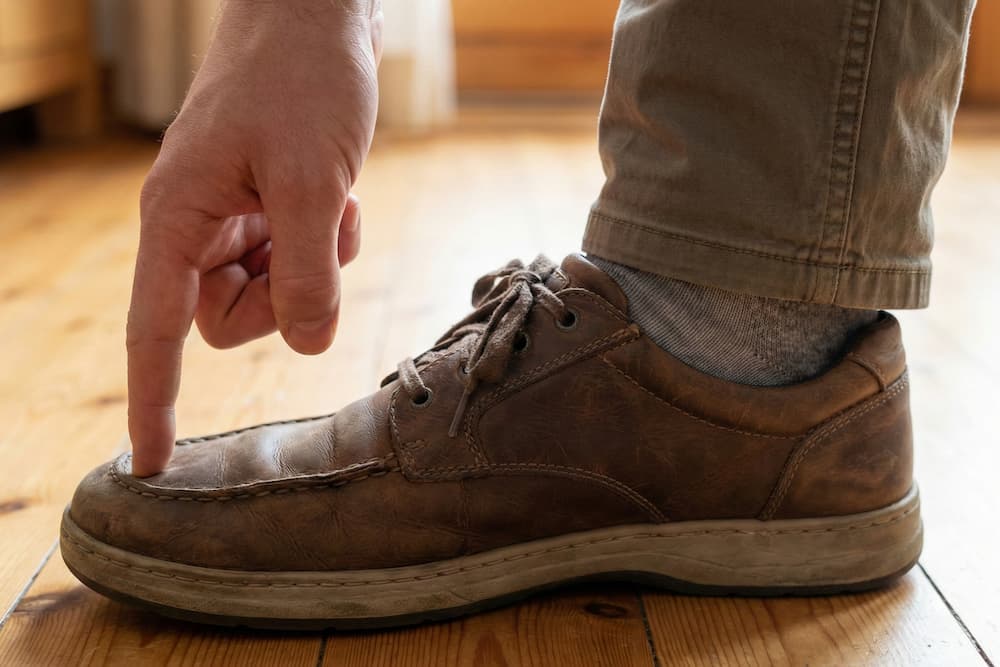 Et nærbillede, der viser den klassiske tommelfingerregel for at tjekke skostørrelse. En pegefinger trykker ned på tåboksen af en brun lædersko for at mærke efter, om der er tilstrækkelig plads mellem storetåen og skosnuden.