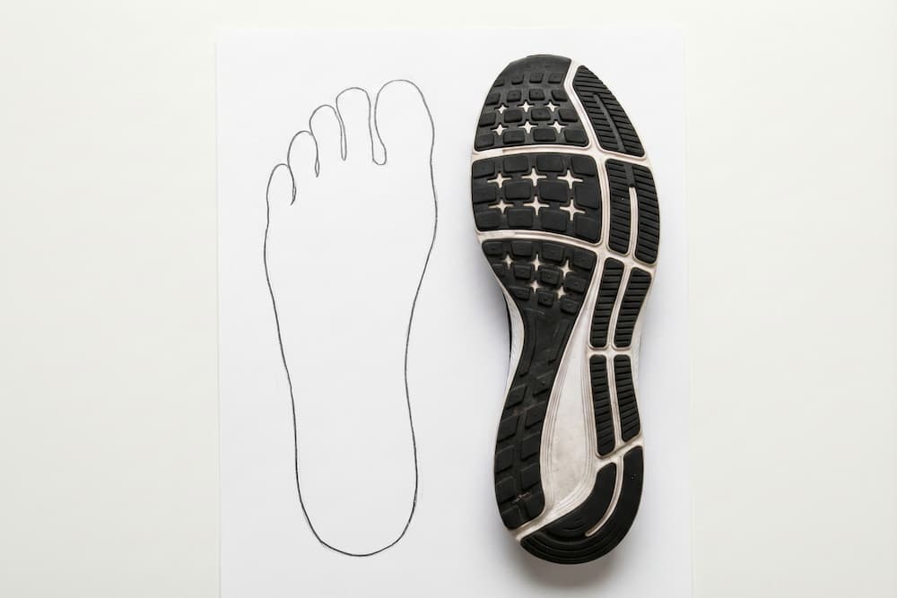 Et 'flat lay' billede, der viser en metode til at tjekke skopasform udefra. Et tegnet omrids af en fod på hvidt papir sammenlignes direkte med undersiden af en løbesko for at se, om sålens form er bred nok til foden.