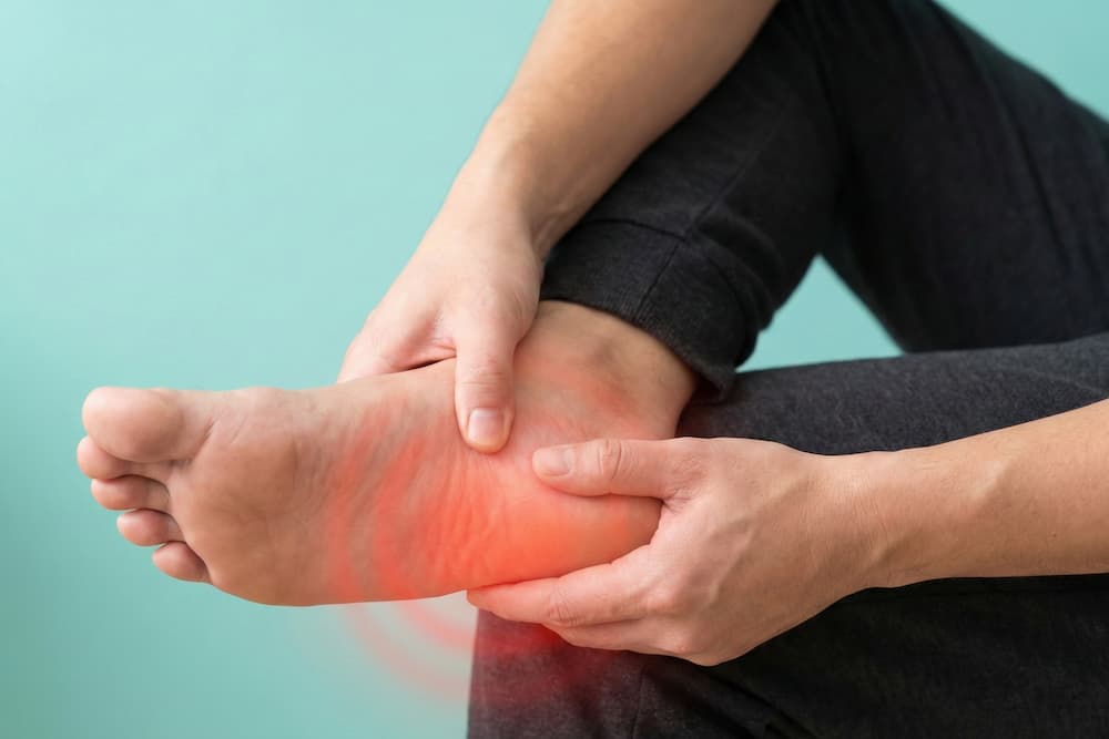 Et nærbillede af en person, der holder om sin fod og masserer hælen med tommelfingeren. Et rødt skær omkring hælen visualiserer smerte og inflammation, hvilket illustrerer symptomerne på almindelige fodlidelser som hælspore eller plantar fasciitis.