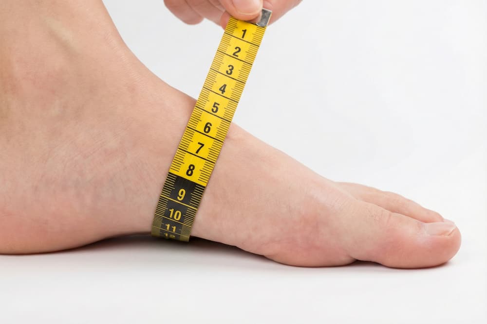 Et nærbillede af en bar fod på en hvid baggrund, hvor vristens højde måles med et gult målebånd. Båndet placeres over fodens højeste punkt for at bestemme fodens volumen, hvilket er afgørende for at vælge sko med den rette dybde og pasform.